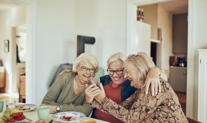 Three older white women are sitting at a table smiling with their arms around each other.