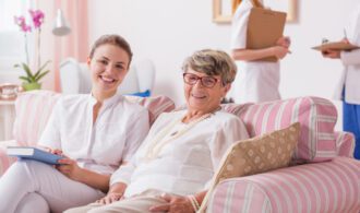 A senior woman and younger woman are in assisted living, smiling and sitting on a couch