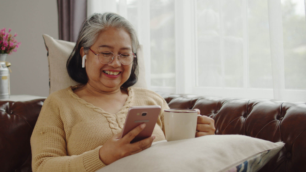 A woman in assisted living sits on a couch with her cell phone and a cup of tea