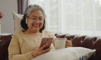 A woman in assisted living sits on a couch with her cell phone and a cup of tea